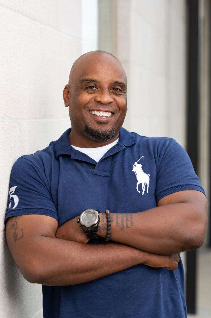 Headshot of Kris leaning against white brick wall.